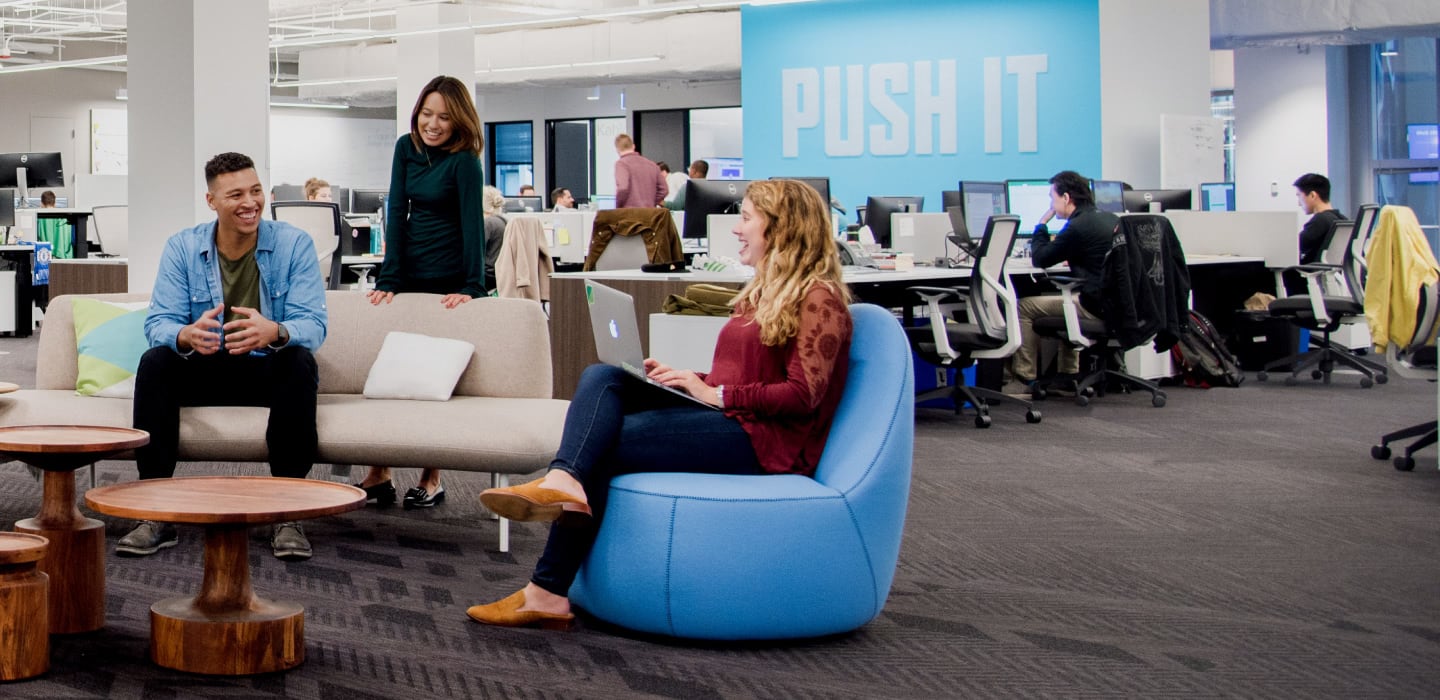 Members of the diverse team at Sprout Social sitting on cool furniture in front of an artwork with the phrase ‘Push It’ made from push pins.